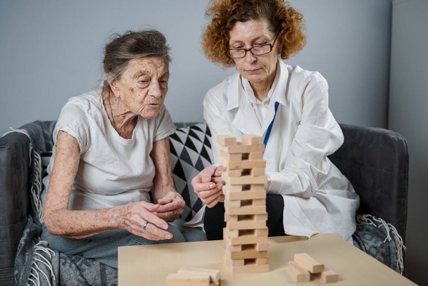 Juego de Jenga. El tema es la demencia, el envejecimiento y los juegos para los ancianos. Mujer mayor caucásica construye torre de bloques de madera con la ayuda de un médico como parte de una terapia y un juego de jenga en el hogar de un paciente. - Foto, imagen
