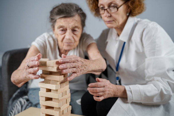 Jenga-Spiel. Themen sind Demenz, Altern und Spiele für alte Menschen. Kaukasische Seniorin baut Turm aus Holzklötzen mit Hilfe eines Arztes im Rahmen eines Therapie- und Jenga-Spiels bei einem Patienten zu Hause. - Foto, Bild