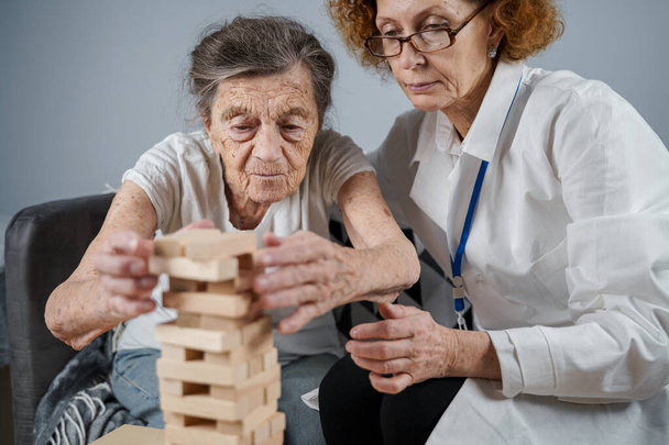 Mature doctor conducting session, therapy for senior patient in nursing home, training fine motor skills for dementia, alzheimer disease and recovery institute by folding wooden blocks, playing jenga. - Photo, Image