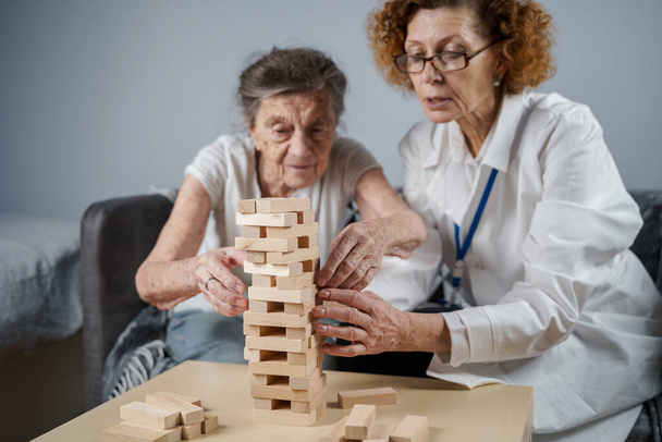Idősebb nő gyakorolni készségek, építeni fából készült blokkok, építeni torony és próbálja meg, hogy ne hagyja leesni, Jenga játék. Idős beteg húzza ki blokk, hely a tetején, támogató orvos a terápia demencia a házban. - Fotó, kép