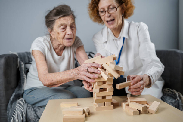 Idősebb nő gyakorolni készségek, építeni fából készült blokkok, építeni torony és próbálja meg, hogy ne hagyja leesni, Jenga játék. Idős beteg húzza ki blokk, hely a tetején, támogató orvos a terápia demencia a házban. - Fotó, kép