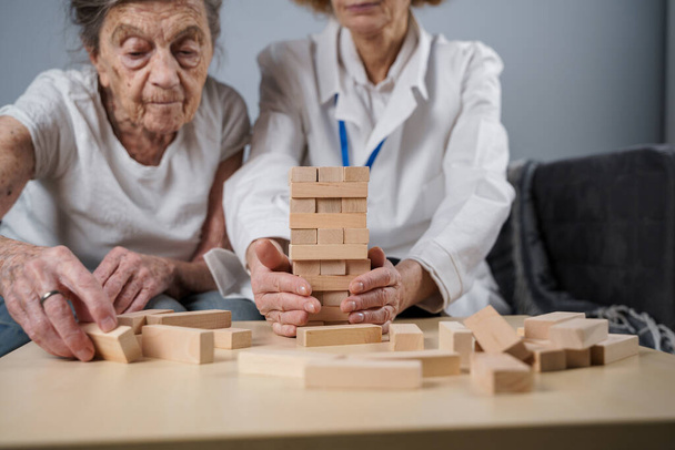 Старша жінка практикує навички, будує дерев'яні блоки, будує вежу і намагається не дати їй впасти, гра в Дженгу. Старий пацієнт витягує блок, зверху, підтримує лікаря під час терапії деменції вдома.. - Фото, зображення