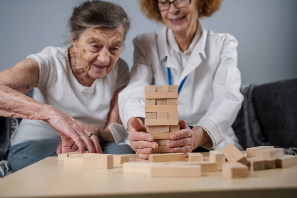 Dementia therapy in playful way, training fingers and fine motor skills, build wooden blocks into tower, playing Jenga. Senior woman 90 years old and doctor playing educational game in nursing home. - Photo, image