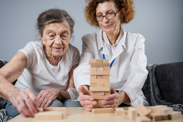 Starsza kobieta ćwiczyć umiejętności, budować drewniane bloki, budować wieżę i starać się nie pozwól mu spaść, gra Jenga. Stary pacjent wyciągnąć blok, miejsce na górze, wsparcie lekarza podczas leczenia demencji w domu. - Zdjęcie, obraz