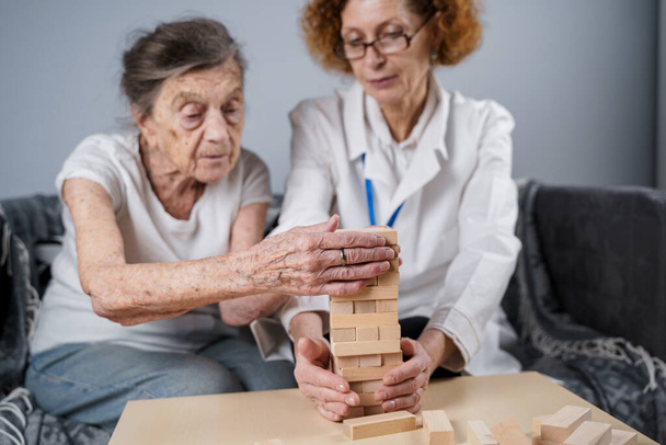 Ανώτερη γυναίκα που παίζει Jenga, χτίζουν πύργο από μπλοκ. Ηλικιωμένος γιατρός σε λευκό εργαστηριακό παλτό, υποστήριξη ανώτερος ασθενής, ανάπτυξη της λογικής, πρόστιμο κινητικές δεξιότητες, Αλτσχάιμερ στο σπίτι με παλιά μοναχική γυναίκα. - Φωτογραφία, εικόνα