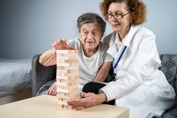 Juego de Jenga. El tema es la demencia, el envejecimiento y los juegos para los ancianos. Mujer mayor caucásica construye torre de bloques de madera con la ayuda de un médico como parte de una terapia y un juego de jenga en el hogar de un paciente. - Foto, Imagen