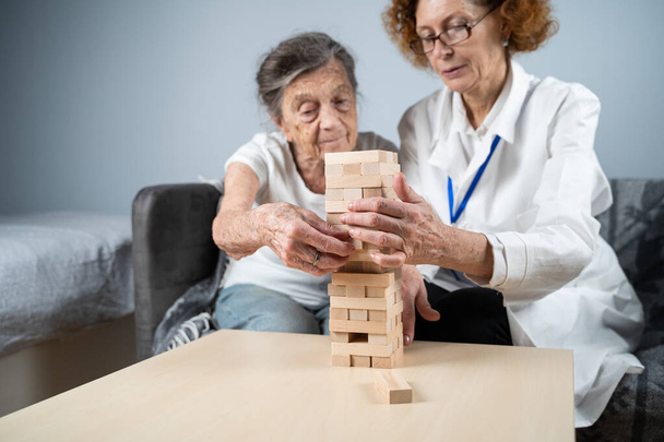 シニア女性の練習スキルは、木製のブロックを構築し、塔を構築し、それが秋にせずにしようとすると、 Jengaゲーム。古い患者は、ブロックを引き出します,上の場所,家の中で治療認知症の間に医師をサポート. - 写真・画像