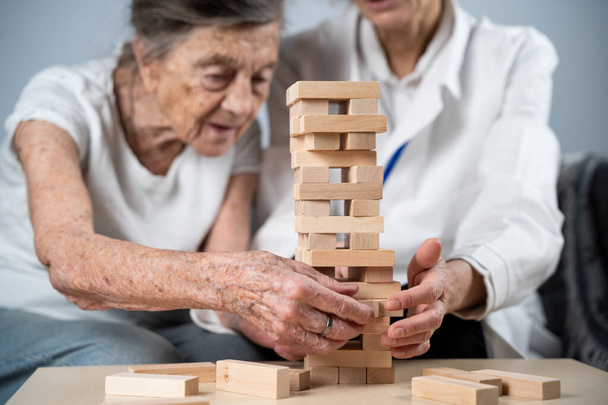 Mature doctor conducting session, therapy for senior patient in nursing home, training fine motor skills for dementia, alzheimer disease and recovery institute by folding wooden blocks, playing jenga. - Photo, Image