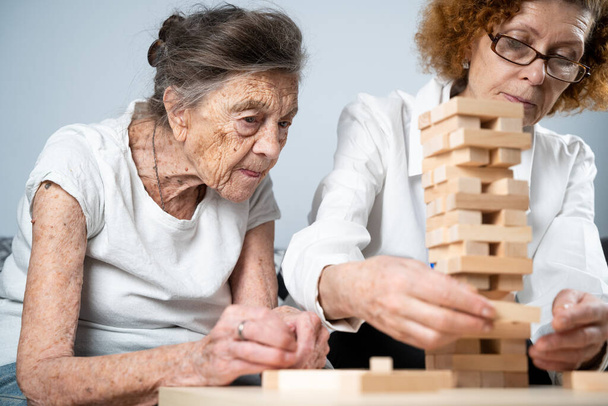 遊び心のある方法で認知症治療は、指や細かい運動能力を訓練し、塔に木製のブロックを構築し、 Jengaを再生します。老人ホームで教育ゲームをプレイするシニア女性90歳と医師. - 写真・画像