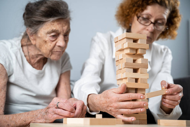 Jenga spel. Thema is dementie, veroudering en spelletjes voor ouderen. Blanke oudere vrouw bouwt toren van houten blokken met de hulp van een arts als onderdeel van een therapie en jenga spel bij een patiënt thuis. - Foto, afbeelding