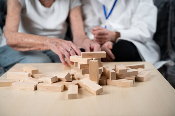 Παιχνίδι Τζένγκα. Θέμα είναι η άνοια, η γήρανση και τα παιχνίδια για τους ηλικιωμένους. Καυκάσια ηλικιωμένη γυναίκα χτίζει πύργο από ξύλινα μπλοκ με τη βοήθεια ενός γιατρού ως μέρος ενός παιχνιδιού θεραπείας και Jenga στο σπίτι ενός ασθενούς. - Φωτογραφία, εικόνα