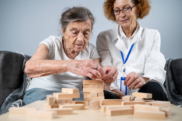Jenga-peli. Teemana on dementia, ikääntyminen ja pelit vanhuksille. Valkoihoinen vanhempi nainen rakentaa torni puiset lohkot avulla lääkäri osana hoitoa ja jenga peli potilaan kotona. - Valokuva, kuva