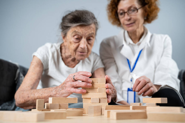 Senior woman practice skills, build wooden blocks, build tower and try not to let it fall, Jenga game. Old patient pull out block, place on top, support doctor during therapy dementia in house. - Foto, Bild