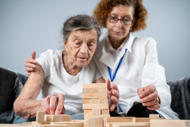 Dementia therapy in playful way, training fingers and fine motor skills, build wooden blocks into tower, playing Jenga. Senior woman 90 years old and doctor playing educational game in nursing home. - Photo, Image