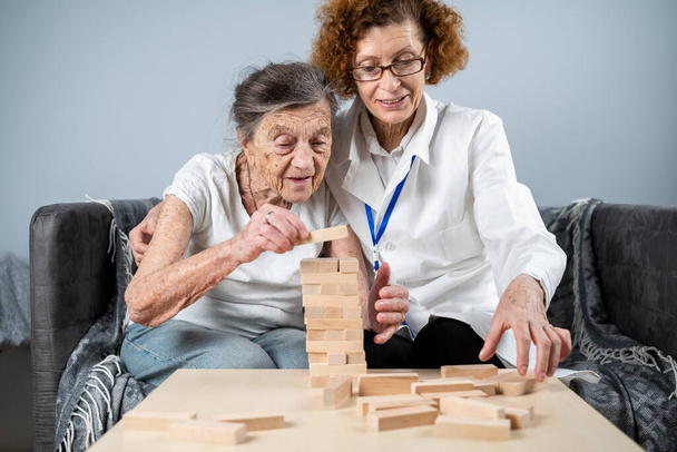 Jenga spel. Thema is dementie, veroudering en spelletjes voor ouderen. Blanke oudere vrouw bouwt toren van houten blokken met de hulp van een arts als onderdeel van een therapie en jenga spel bij een patiënt thuis. - Foto, afbeelding