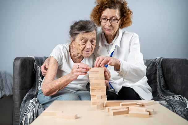 Senior Frau Jenga spielen, bauen Turm aus Blöcken. Älterer Arzt im weißen Laborkittel, unterstützt ältere Patientin, entwickelt Logik, Feinmotorik, Alzheimer zu Hause bei alten einsamen Frauen. - Foto, Bild