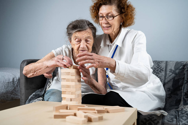 Παιχνίδι Τζένγκα. Θέμα είναι η άνοια, η γήρανση και τα παιχνίδια για τους ηλικιωμένους. Καυκάσια ηλικιωμένη γυναίκα χτίζει πύργο από ξύλινα μπλοκ με τη βοήθεια ενός γιατρού ως μέρος ενός παιχνιδιού θεραπείας και Jenga στο σπίτι ενός ασθενούς. - Φωτογραφία, εικόνα