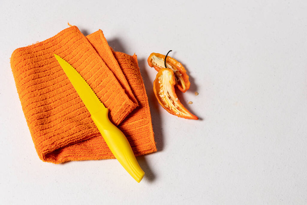 Pano de prato laranja dobrado com uma pequena faca amarela ao lado de uma pimenta no topo em um fundo branco - Foto, Imagem