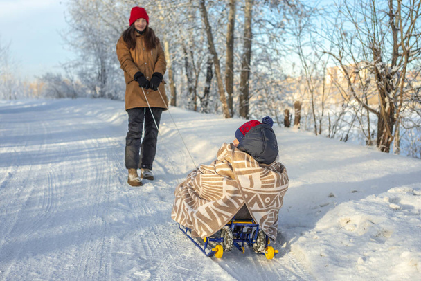 Mom is pulling a child on a sled walking on a frosty winter Sunny day out of doors. Baby wrapped in a blanket rides on a sledge - Photo, image