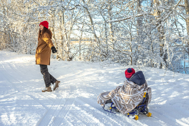 Mom is pulling a child on a sled walking on a frosty winter Sunny day out of doors. Baby wrapped in a blanket rides on a sledge - Foto, Imagen