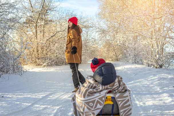Mama zieht ein Kind auf einem Schlitten, der an einem frostigen Wintersonntag draußen vor der Tür steht. Baby in Decke gehüllt fährt auf Schlitten - Foto, Bild