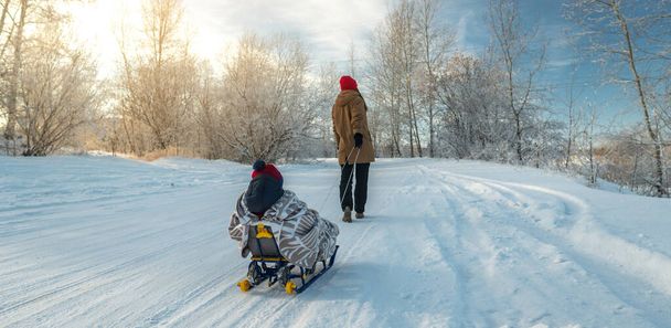 A mother with a child walks on a frosty winter morning on a Sunny day. Baby wrapped in a blanket rides on a sled - Φωτογραφία, εικόνα