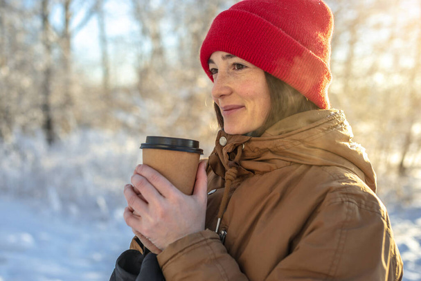 Молодая женщина пьет горячий кофе в морозный зимний солнечный день на открытом воздухе в лесу - Фото, изображение