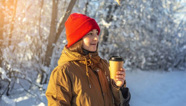 Молодая женщина пьет горячий кофе в морозный зимний солнечный день на открытом воздухе в лесу - Фото, изображение