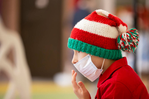 A child in a medical mask wearing a Santa Claus hat due to the coronavirus pandemic. Sad little boy wearing a Christmas hat during the coronavirus epidemic. - Photo, Image