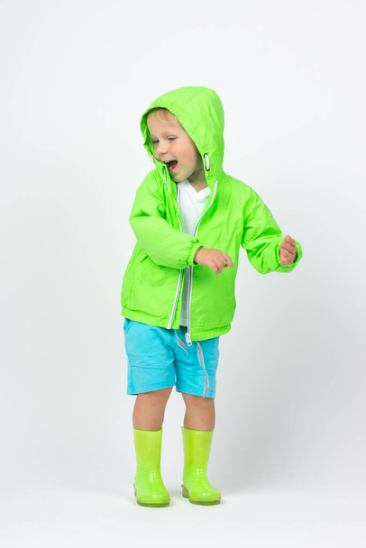明るいライムグリーンのジャケットとラバーブーツを着た子供の男の子が笑い、白い背景で踊ります。悪天候、水たまりや湿気のための春の服についての垂直ショットの概念. - 写真・画像