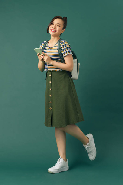 Ελκυστική χαρούμενη κυρία influencer κρατήσει τηλέφωνο φορώντας σακίδιο στους ώμους σε πράσινο φόντο - Φωτογραφία, εικόνα