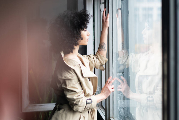 jonge mooie vrouw staat in de buurt van het raam in een regenjas - Foto, afbeelding