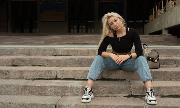 Stylish young blonde woman in jeans sitting outdoors on long stone stairs - Fotó, kép