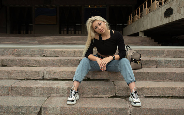 Stilvolle junge blonde Frau in Jeans sitzt draußen auf der Treppe - Foto, Bild