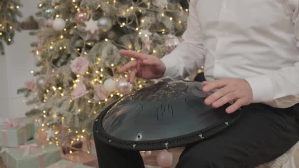 Un uomo gioca appeso nella stanza con decorazioni natalizie. musicista suona il tamburo serbatoio. Musicista suona il handpan. Musicista suona il tamburo lingua in acciaio - Filmati, video