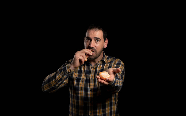 Bearded man dressed in yellow plaid shirt offering an orange with one hand and eating a slice with the other hand on black studio background. Healthy food concept - Photo, Image
