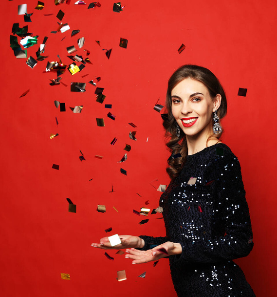 Holiday and party concept: beautiful young woman wearing evening dress standing under confetti rain over red background. Happy time. - Valokuva, kuva