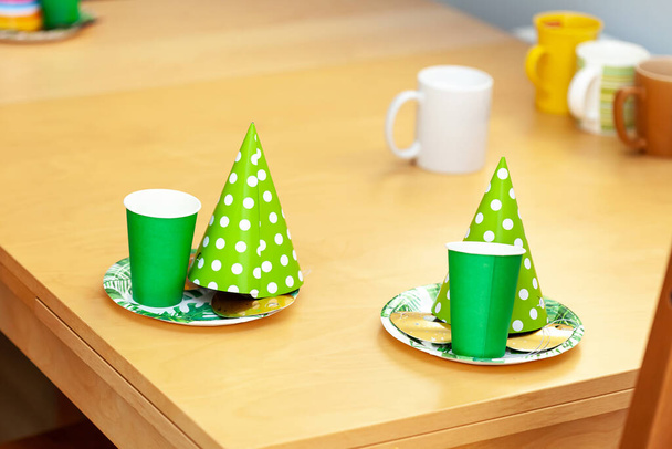 Sombreros de fiesta, platos de papel, máscaras y copas verdes colocados en la mesa listos para una fiesta de cumpleaños de los niños, dos juegos, objetos, primer plano, nadie. Celebración de cumpleaños de niños simples en el concepto de preparación para el hogar - Foto, Imagen