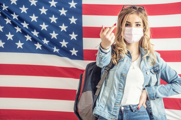 Young blonde female student shows middle finger gesture to the camera while standing in front of the US flag. - Fotografie, Obrázek