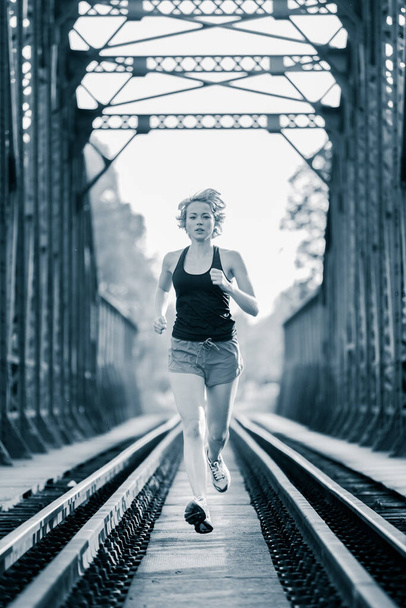Athlete running on railaway bridge training for marathon and fitness. Healthy sporty caucasian woman exercising in urban environment before going to work. Active urban lifestyle - Photo, Image