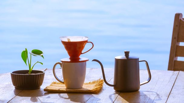 Damla kahve fincanı seti, çiçek saksısında yeşil bitki, ahşap yuvarlak masa, nehir kenarı manzarasına karşı sabah vakti. - Fotoğraf, Görsel