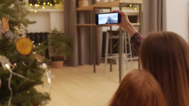 Mladá žena a její dcera s video hovor s mužem v venkovní zimní lokalitě, zatímco sedí v útulném obývacím pokoji vedle krásné zdobené vánoční stromeček - Záběry, video
