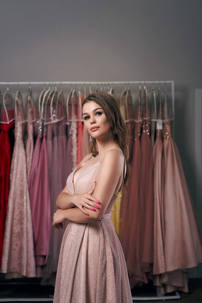 Portrait of young beautiful girl wearing a full-length pale pink glitter chiffon draped prom ball gown. Model selecting an outfit for occasion in dress hire service with many options on background. - Fotó, kép
