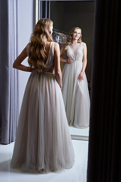 Young beautiful blonde girl wearing a full-length silver white chiffon prom ball gown decorated with sparkles and sequins. Model in front of mirror in a fitting room at dress hire service. - Foto, Imagem