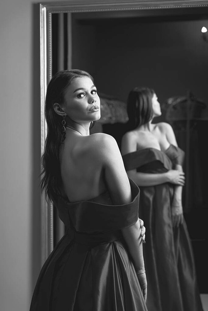 Young beautiful girl wearing an off-the-shoulder full-length satin slit prom ball gown. Model looking in mirror. Black and white image in retro style. - Fotografie, Obrázek