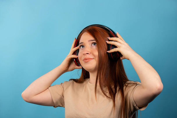 Authentic redhaired girl with big headphones, listens to music and looks up. Isolated on a blue background. High quality photo - Photo, Image
