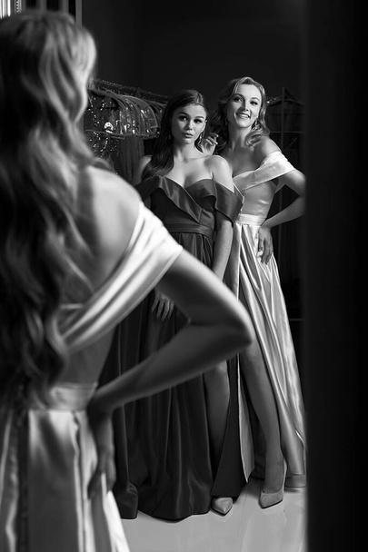 Two young beautiful girls wearing off-the-shoulder full-length sky blue and crimson red satin slit prom ball gowns. Models looking in mirror. Fitting room in dress hire service. - Foto, imagen