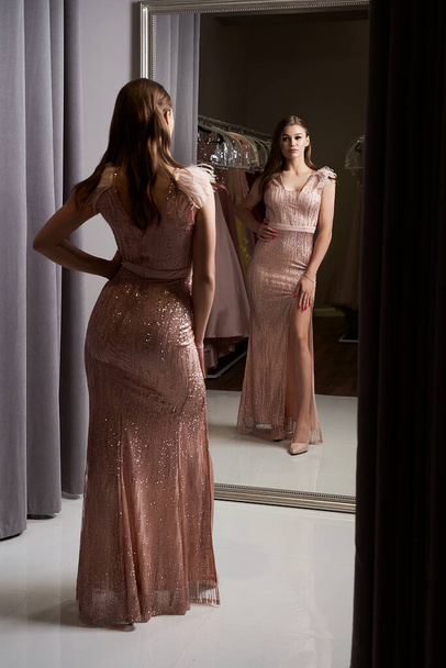 Young beautiful brunette girl wearing a full-length pale pink fitting chiffon prom ball slit gown decorated with sparkles and sequins. Model in front of mirror in a fitting room. - Foto, Imagem