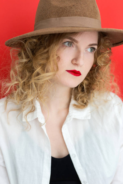 Young blonde woman with green eyes wearing in hat and white shirt over red background. Close up portrait of student curly girl. Beauty concept. - Foto, afbeelding