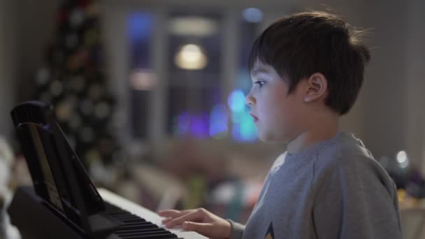 Niño tocando el piano, Niño aprendiendo música con un piano eléctrico en la sala de estar, Niño relajándose cantando una canción en casa en la noche de Navidad - Metraje, vídeo